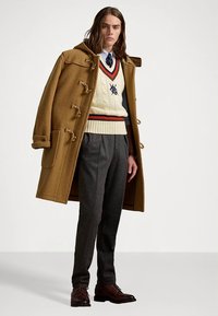 Καφετί μάλλινο παλτό duffle με κούμπωμα με κουμπιά, εκρού πουλόβερ με πλεχτό σχέδιο και κόκκινες και μπλε ρίγες, γκρι παντελόνι και καφέ δερμάτινα παπούτσια.