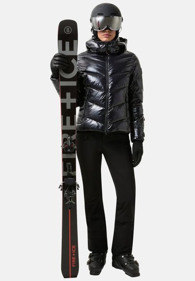 Zwarte gewatteerde jas met een glanzende afwerking, zwarte skibroek, zwarte handschoenen en zwarte skischoenen. Houdt zwarte ski's met rode accenten vast.