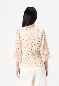 Mujer con cabello largo y oscuro vestida con una blusa crema con estampado floral rojo y mangas plisadas, combinada con pantalones blancos texturizados, vista desde atrás.