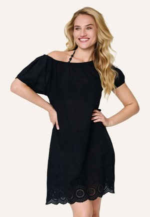 Frau mit langen blonden Haaren, die lächelt und ein kurzes schwarzes, schulterfreies Kleid mit gewelltem Spitzenabschluss trägt, Hände in die Hüften gestützt.