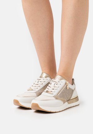 Zapatos deportivos blancos y dorados con paneles laterales de tela texturizada, cordones, cremalleras metálicas y un collar acolchado. Suela de goma con acentos marrón claro.