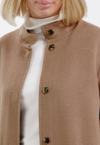 Hellbrauner, strukturierter Mantel mit einem gewellten Kragen und drei Knöpfen. Über einem weißen Rollkragenpullover getragen, der aus glattem Fabric besteht und eine taillierte Passform hat.