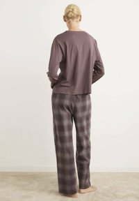Camicia marrone a maniche lunghe con vestibilità rilassata, abbinata a pantaloni da pigiama in tartan nei toni di viola, grigio e bianco. Tessuto morbido.