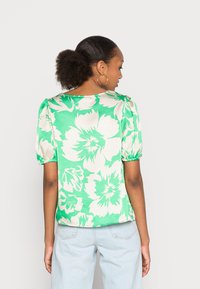 Blusa verde de manga curta com padrões grandes de flores brancas, com um corte solto e mangas bufantes franzidas, combinada com jeans azul claro.