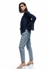 Blusa de gasa azul marino con detalles de volantes, de manga larga. Combinada con unos vaqueros de mezclilla azul claro y sandalias blancas con tiras.