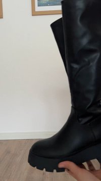 Svart knä-hög stövel gjord av slät läder med en kraftig sula och minimal detaljering. Har en lätt glans och tjock, texturerad sula.