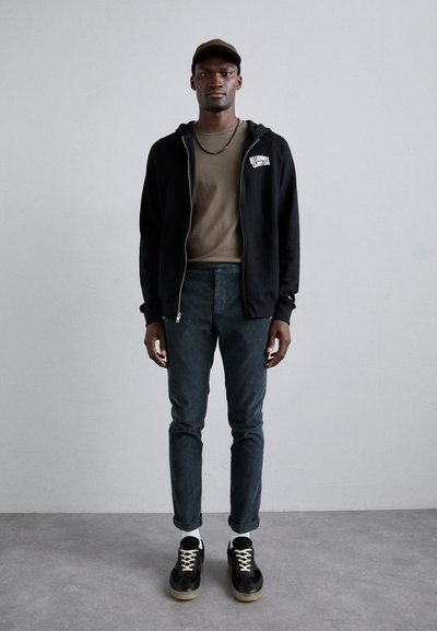 Sort zip-up hættetrøje, olivengrøn T-shirt, mørkegrønne bukser og sorte sneakers med lyse såler. Enkel design, ingen synlige mønstre.
