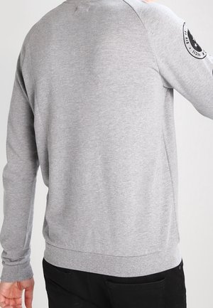 Sudadera gris de manga larga, con puños y dobladillo acanalados, que presenta un parche con un logotipo circular en la manga izquierda. Textura de tela suave y lisa.