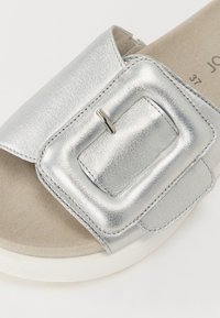 Sandale glissante en métal argenté avec un grand détail de boucle rectangulaire. Semelle intérieure beige douce et semelle extérieure blanche. Texture lisse, design décontracté.