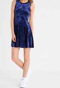 Granatowa aksamitna sukienka o bezrękawnikowym kroju, dopasowanej górze i rozszerzanej spódnicy. W połączeniu z białymi trampkami dla uzyskania swobodnego wyglądu.