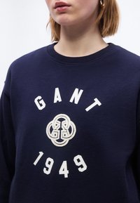 Γυναίκα που φοράει μπλε σκούρο φούτερ με λευκό λογότυπο GANT, διαπλεκόμενο σύμβολο G και το κείμενο "1949" στο μπροστινό μέρος, δείχνοντας την περιοχή του λαιμού και των ώμων.
