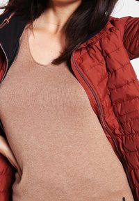Röd pufferjacka med en quiltad textur, med en svart dragkedjedetalj, bärs över en beige ärmlös topp av ribbat material.