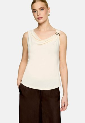 Femme portant un haut crème sans manches avec un col drapé et une boucle dorée décorative sur l'épaule gauche, associé à un pantalon brun foncé.