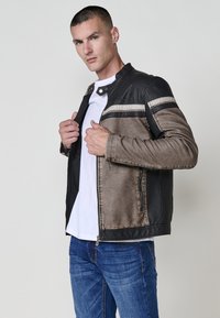 Uomo che sistema la cerniera di una giacca in pelle nera e marrone indossata sopra una camicia bianca e jeans blu, su uno sfondo leggero e uniforme.