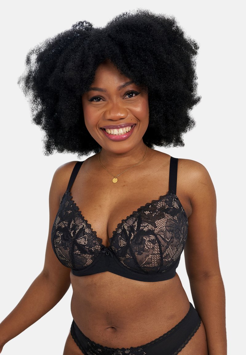 Smiling woman with natural curly hair wearing a black lace bra and matching underwear on a plain white background.
