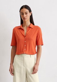 Korte mouwen, kraagloze oranje blouse met knoopsluiting, gemaakt van lichtgewicht stof. Gepaard met lichtbeige op maat gemaakte broeken.