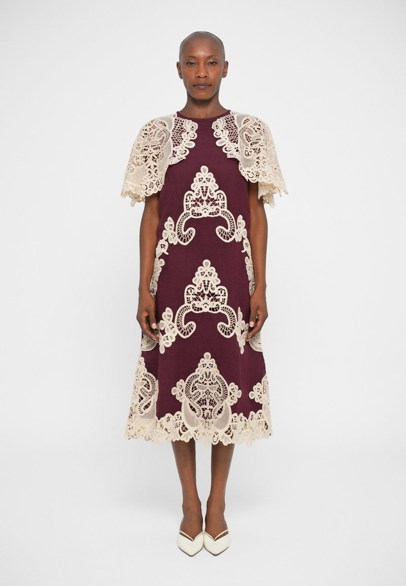 Vestido bordeaux com detalhes em renda creme, apresentando padrões intrincados e uma bainha em forma de onda. Sobreposição de capa de renda curta nos ombros.