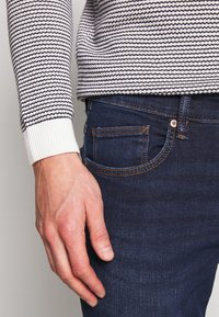 Randig stickad tröja med en ribbad vit mudd, kombinerad med mörkblå denimjeans som har en ficka med orange sömnad.
