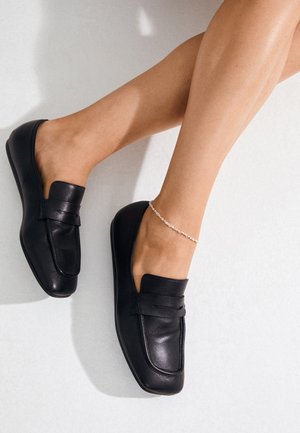 Legs wearing black leather loafers and a delicate silver anklet resting on a light surface.
