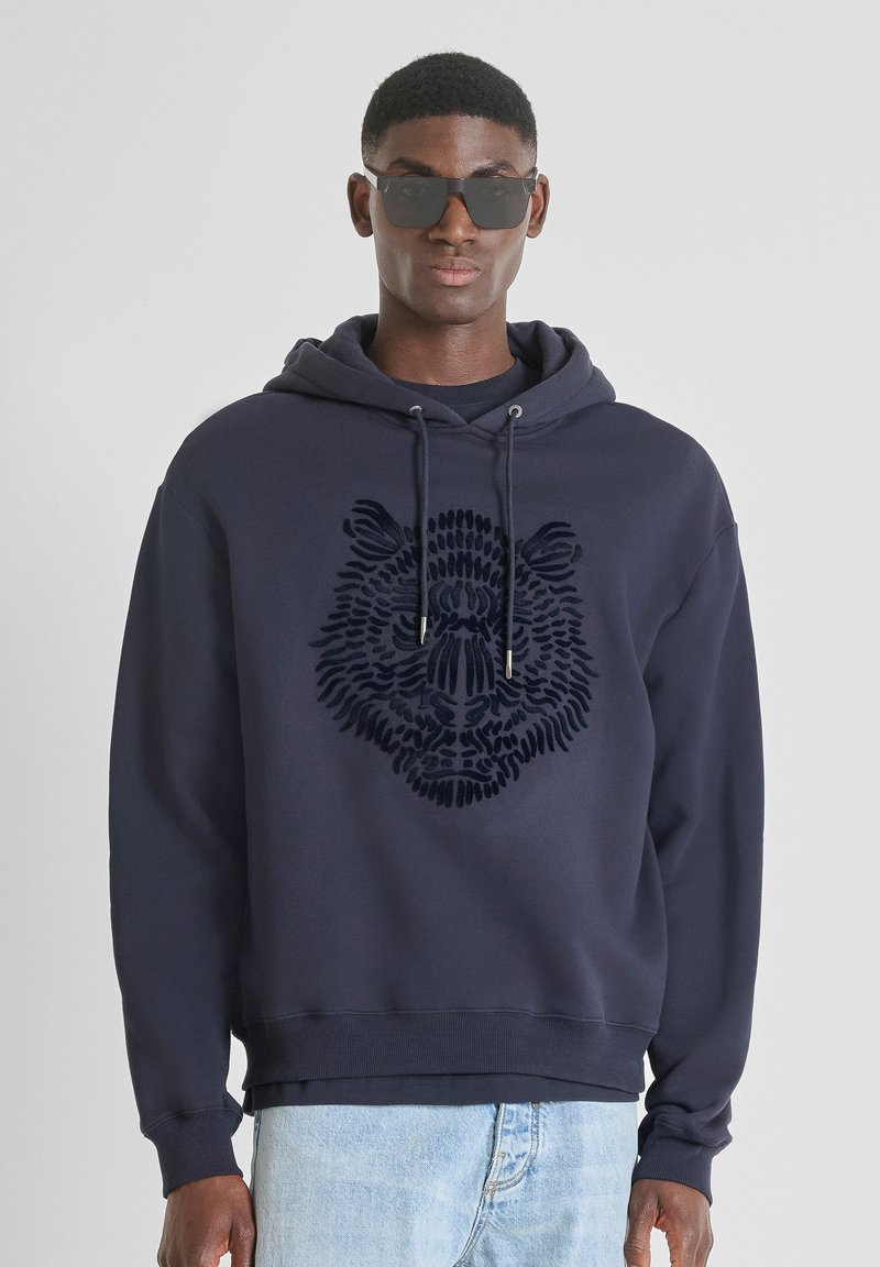 Felpa blu marina con un design di un volto di leone testurizzato. Presenta un cappuccio, cordini e polsini a costine. Indossata con jeans in denim chiaro e occhiali da sole.