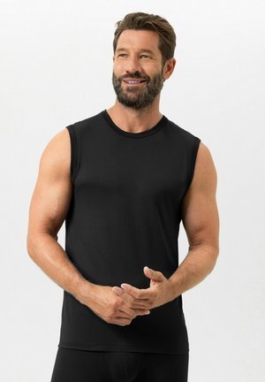 Muskulöser Mann mit Bart, der ein schwarzes ärmelloses Hemd trägt, steht mit verschränkten Händen, schaut leicht zur Seite und lächelt vor weißem Hintergrund.