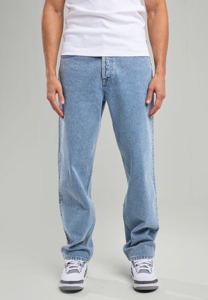 Jean en denim bleu clair avec une coupe décontractée, design à cinq poches et un léger délavage. Associé à des baskets blanches avec des accents gris.