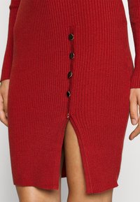 Robe en tricot côtelé rouge avec une fente montée jusqu'à la cuisse et cinq boutons noirs sur le côté pour la fermeture et le détail de conception.