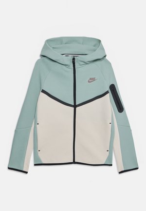Giacca con cappuccio in azzurro chiaro e crema. Dotata di zip frontale, accenti neri a contrasto e una tasca con zip sulla manica per riporre oggetti.