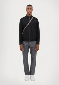 Črna jakna z zadrgo in stoječim ovratnikom, sive jeans hlače, ter bele superge. Model stoji pokončno in prikazuje prileganje ter dizajn obleke.