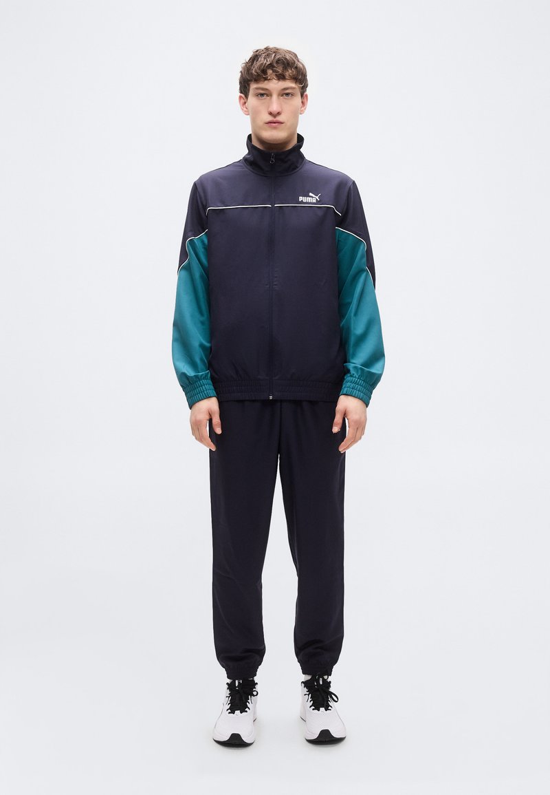 Σκούρο μπλε φόρμα με αντιθετικές πράσινες μανσέτες, μπουφάν με φερμουάρ, ελαστικές μανσέτες και παντελόνι που ταιριάζει. Φορεμένο με λευκά παπούτσια.