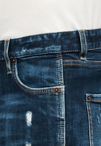 Gros plan sur un jean en denim bleu usé avec des coutures orange et de petits endroits usés, associé à un haut blanc côtelé.