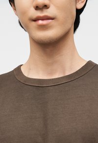 Νεαρό άτομο με κοντά σκούρα μαλλιά που φορά καφέ μπλούζα με στρογγυλή λαιμόκοψη, δείχνοντας το κάτω μέρος του προσώπου, το λαιμό και το πάνω μέρος του στήθους με φόντο μονόχρωμο.