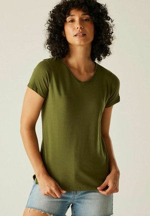 Mujer con cabello rizado que lleva una camiseta verde oliva de manga corta y cuello en V y pantalones cortos vaqueros azul claro, de pie contra un fondo liso y claro.