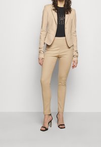 Mujer vestida con un blazer y pantalones beiges ajustados, con una camiseta gráfica negra y sandalias de tacón negro, de pie contra un fondo liso.