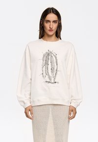 Sudadera color crema con una ilustración de un cactus en gris y negro. Presenta un corte relajado y puños acanalados. Usada sobre una falda transparente y texturizada.