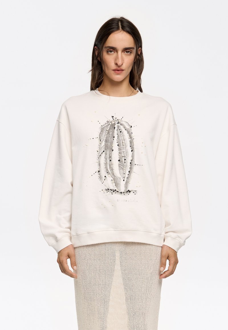 Sudadera color crema con una ilustración de un cactus en gris y negro. Presenta un corte relajado y puños acanalados. Usada sobre una falda transparente y texturizada.