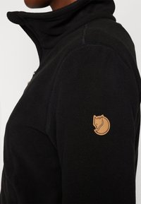 Svart fleecejacka med hög krage, texturerad tyg och en tan-färgad cirkulär logotyp med en rävdesign på vänster axel.