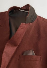 Blazer homme couleur rouille avec doublure intérieure foncée, col marron contrastant et pochette grise dans la poche poitrine.
