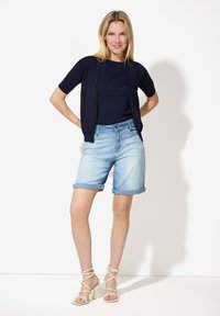 Navy Kurzarm-Cardigan über einem navy-blauen Top, kombiniert mit hellblauen Denim-Shorts und beigen Riemchensandalen. Sanfte Texturen und lässiges Design.