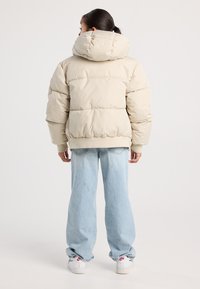 Persona di spalle che indossa un giubbotto imbottito beige con cappuccio, jeans svasati blu chiaro e sneakers bianche Nike con loghi rossi su uno sfondo bianco.