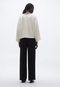 Krém színű, túlméretezett pulóver nyers szegéllyel, magas derekú, fekete, bő szárú nadrággal párosítva. Simaságú anyag, minimalista kialakítás.