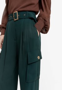 Tyrkysové široké cargo kalhoty s hlubokými bočními kapsami, zlatými detaily a ladícím páskem; vyrobené z hladkého, lehkého materiálu.