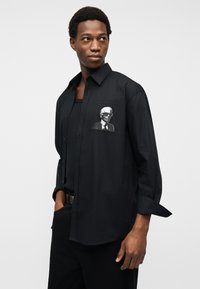 Άνδρας που φοράει μαύρο πουκάμισο με κουμπιά και λευκό γραφικό ενός άνδρα με γυαλιά ηλίου στην τσέπη του στήθους, στέκεται με το ένα χέρι στην τσέπη.