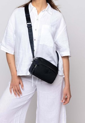 Black crossbody bag made of smooth material, featuring a zip closure, adjustable patterned strap, and embossed logo detail on front.