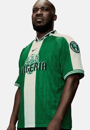 Mężczyzna noszący zielono-biały strój piłkarski Nigerii z narodowym emblematem orła i naszywką Nigeria Football Association na rękawie.