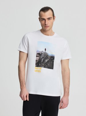 Giovane uomo con capelli pettinati all'indietro che indossa una t-shirt bianca con una foto di una scogliera e la scritta "OUT OF OFFICE", abbinata a pantaloni neri.