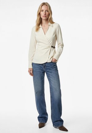 Frau trägt eine cremefarbene, gestreifte Wickelbluse mit Schnalle, blaue Jeans mit weitem Bein und braune spitze Schuhe, steht mit einer Hand in der Tasche.