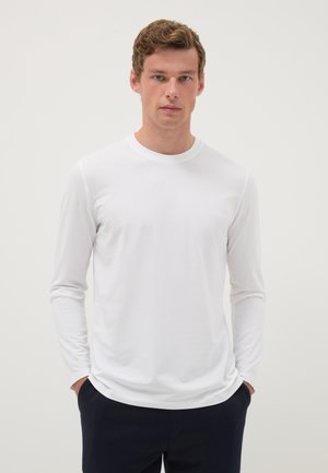 Jonge man met krullend haar, gekleed in een eenvoudig wit shirt met lange mouwen en donkere broek, staand met handen in de zakken tegen een neutrale achtergrond.