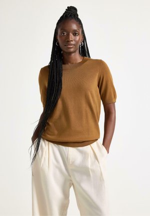 Kvinde med langt, flettet hår iført brun kortærmet sweater og cremefarvede bukser med høj talje, stående med hænderne i lommerne mod en hvid baggrund.