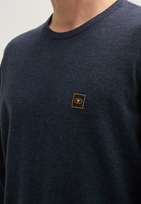 Granatowy sweter dzianinowy z okrągłym dekoltem, z pomarańczowym kwadratowym logo na piersi. Miękka faktura, klasyczny krój.